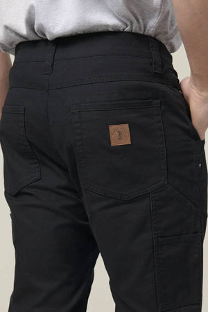 A person wears Hard Yakka Heritage Carpenters Pant, featuring black fabric, visible back pockets, and a brown leather patch—shown waist down with hand on hip—ideal for a rugged workwear look.