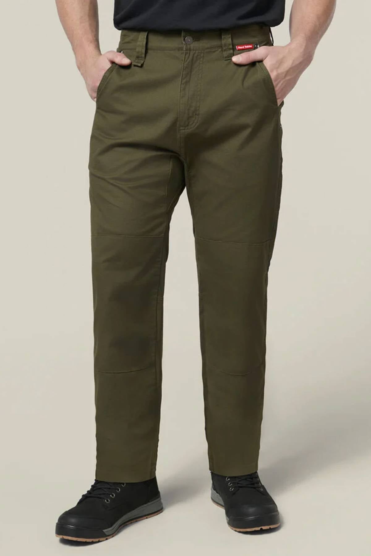 A person wearing olive green Hard Yakka Heritage Carpenters Pant, a black shirt, and black boots stands with hands in pockets against a plain light background. A red tag is visible on the pants near the right pocket.