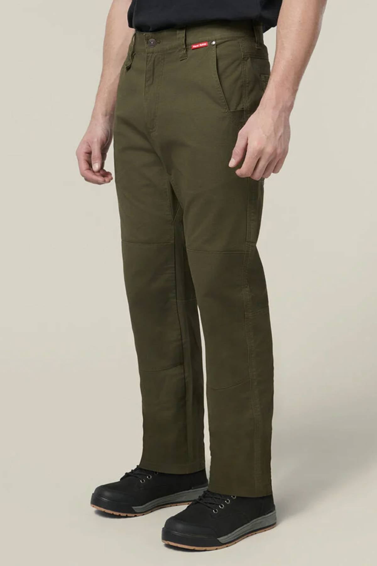 A person in olive green Hard Yakka Heritage Carpenters Pants, black shoes, and a black shirt stands against a plain light background. The relaxed-fit workwear pants feature a small red tag near the pocket.