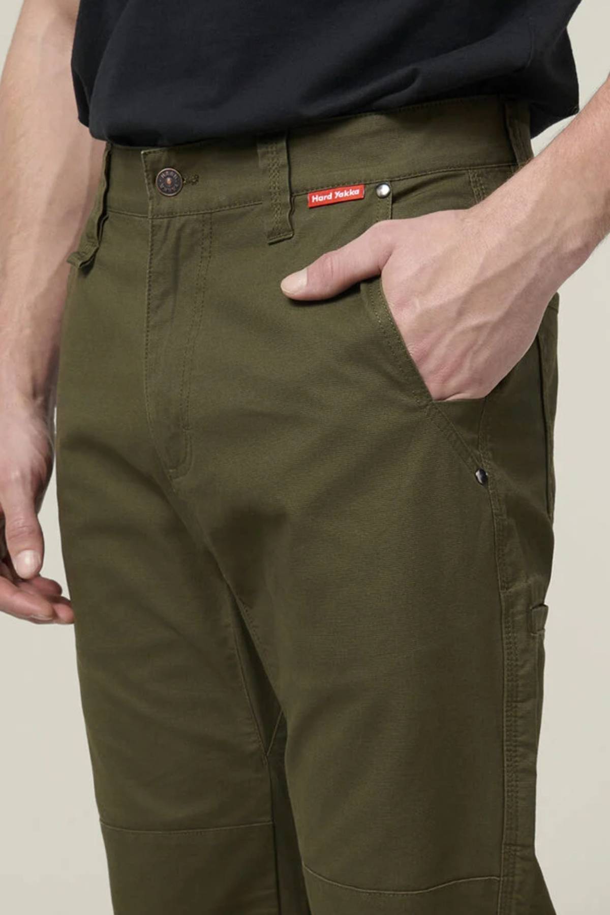 A person wearing Hard Yakka Heritage Carpenters Pant in olive green and a black shirt poses with their right hand in the pocket, featuring a close-up of the front and the signature small red label above the pocket, showcasing classic workwear style.