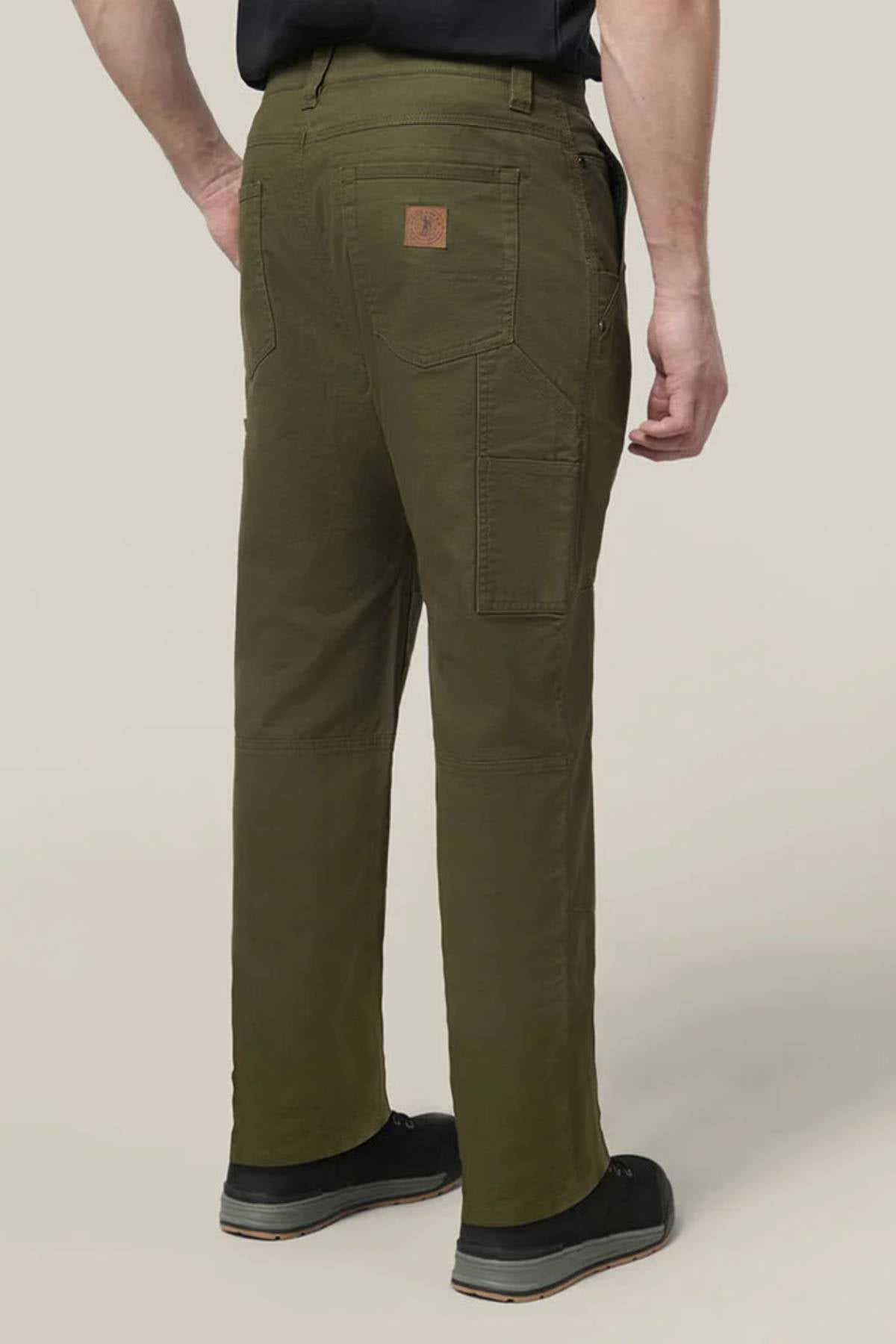 A person in olive Hard Yakka Heritage Carpenters Pant and black shoes stands facing away, hands on hips. The pants have large back pockets and a brown leather patch on the waistband.
