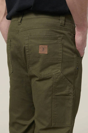 Person wearing olive green Hard Yakka Heritage Carpenters Pant with a brown patch above the back pocket, standing with one hand in their pocket against a plain background—ideal for workwear style.