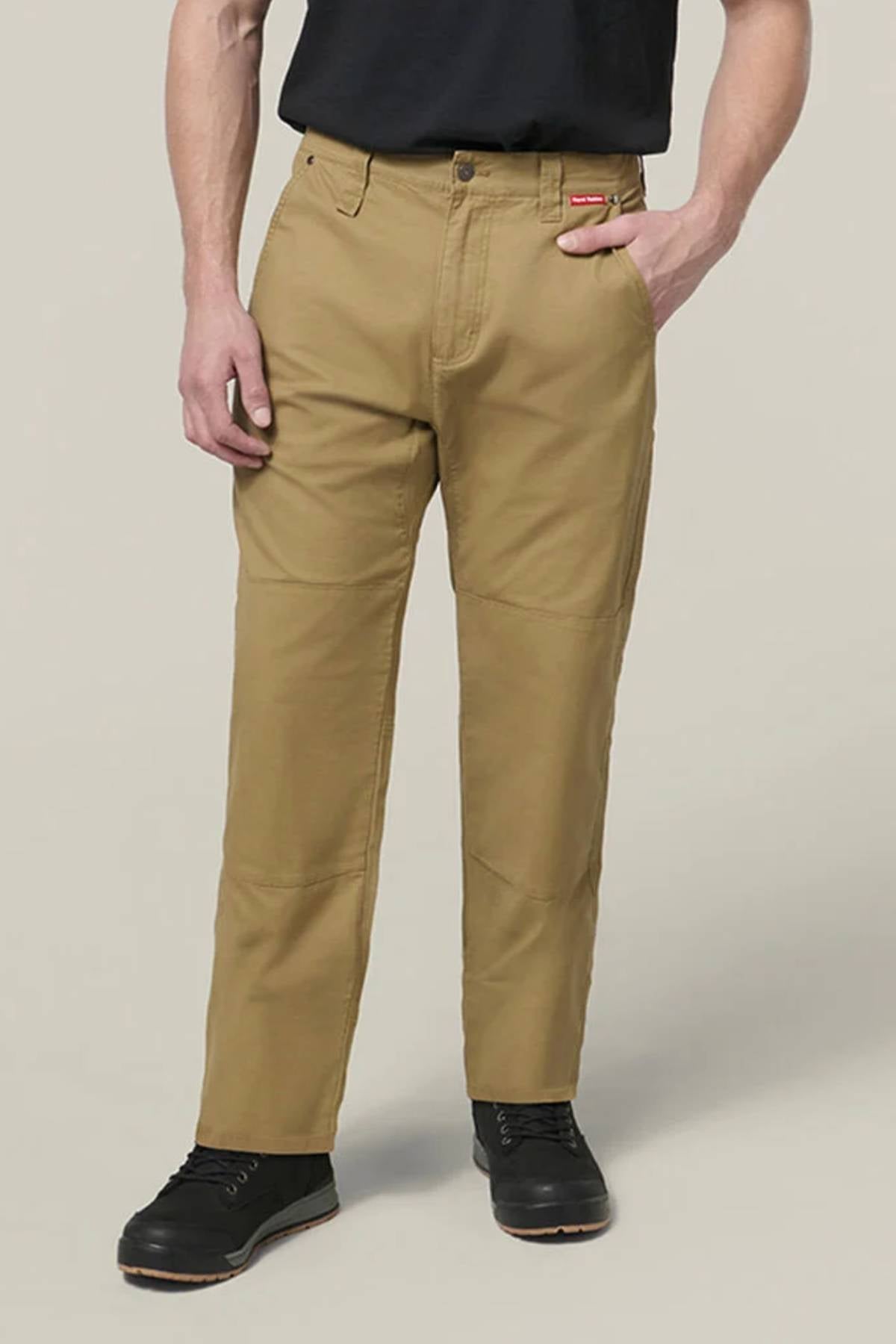 A person in tan Hard Yakka Heritage Carpenters Pant, a black t-shirt, and black work shoes stands with one hand in their pocket against a plain background, capturing iconic workwear style.