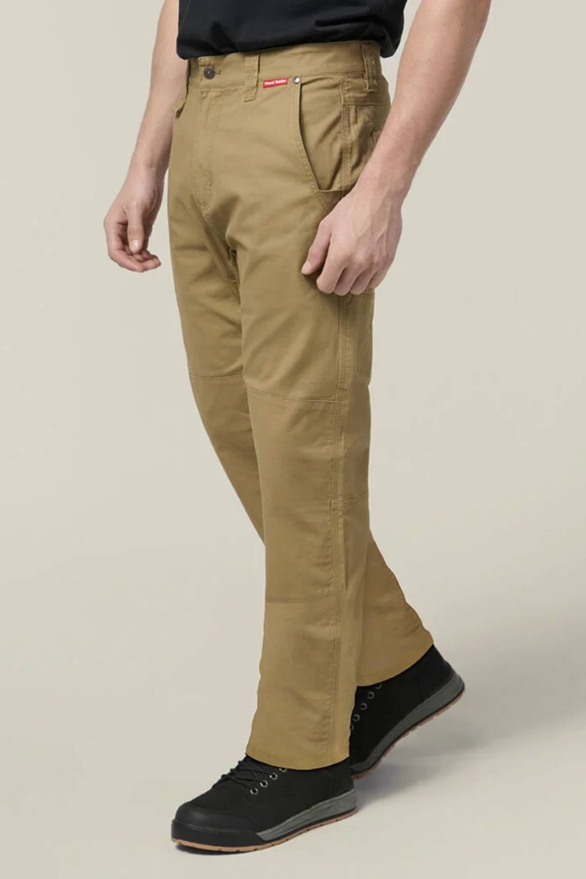 Someone wears tan Hard Yakka Heritage Carpenters Pant with black lace-up shoes, standing against a plain light background, one leg forward and hands relaxed by their sides—a timeless workwear-inspired look.
