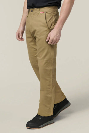 Someone wears tan Hard Yakka Heritage Carpenters Pant with black lace-up shoes, standing against a plain light background, one leg forward and hands relaxed by their sides—a timeless workwear-inspired look.