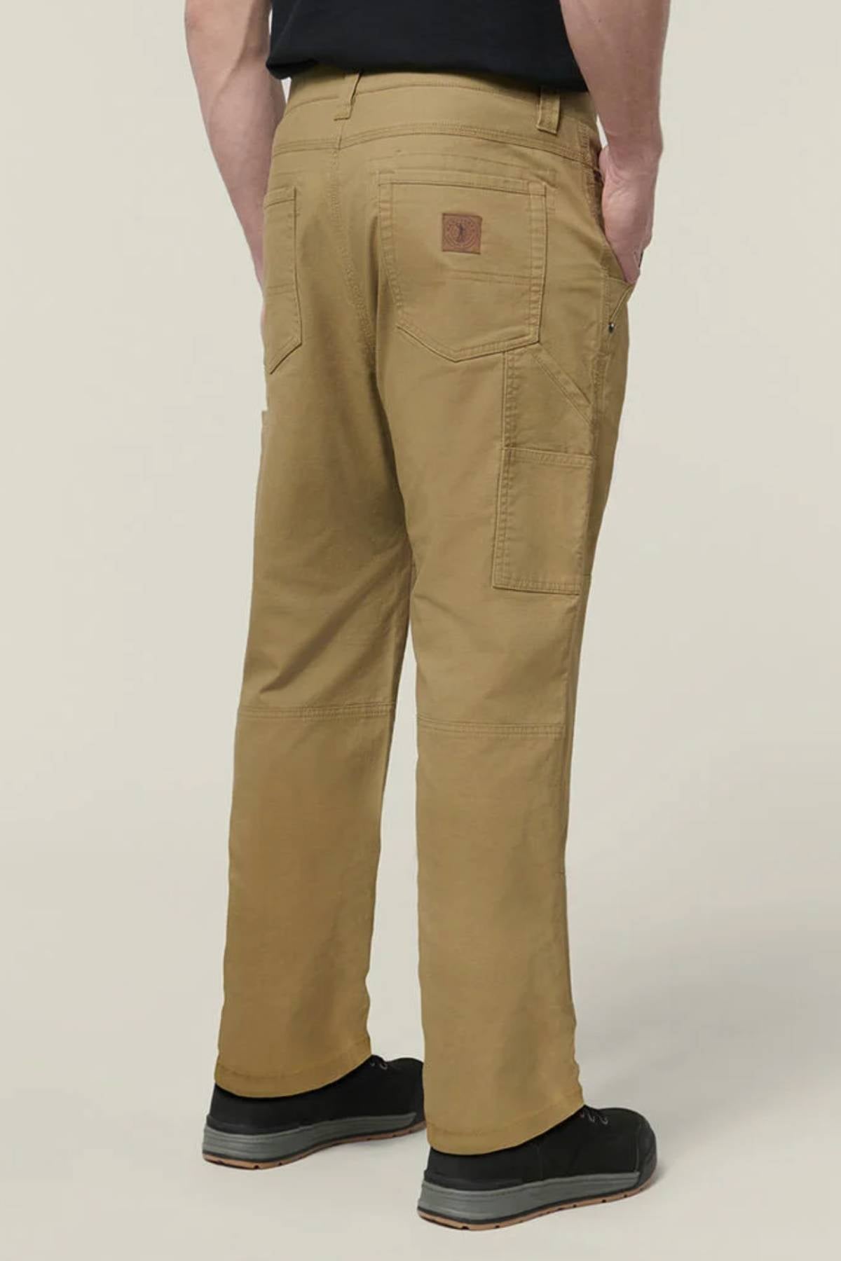 Shown from the back against a plain light background, a person stands with hands in pockets, wearing black shoes and Hard Yakka Heritage Carpenters Pant—demonstrating classic workwear style.