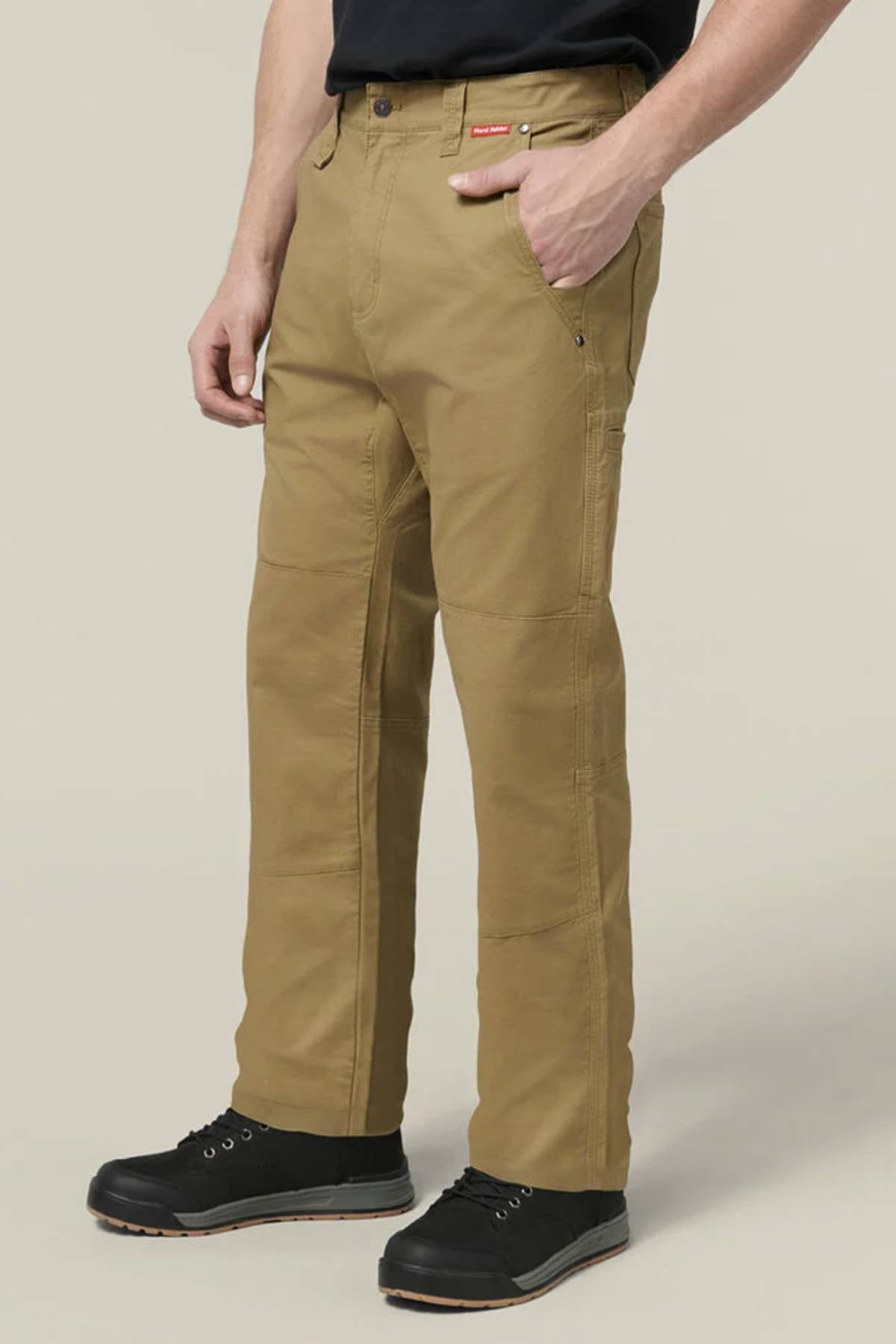 Wearing Hard Yakka Heritage Carpenters Pants, a black t-shirt, and black shoes, a person stands against a plain light background with one hand in their pocket.