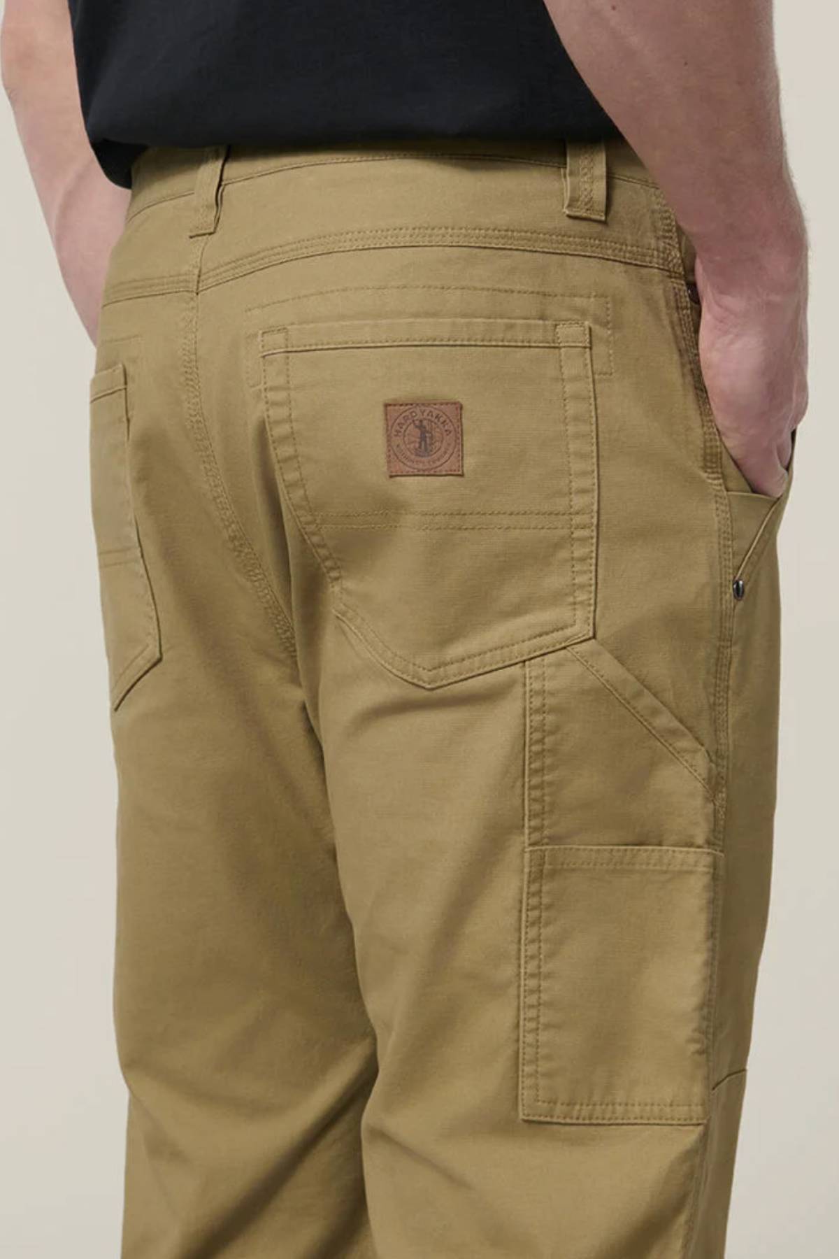 A person stands with hands in pockets wearing Hard Yakka Heritage Carpenters Pant, featuring multiple pockets and a brown leather patch on the back, against a neutral background.