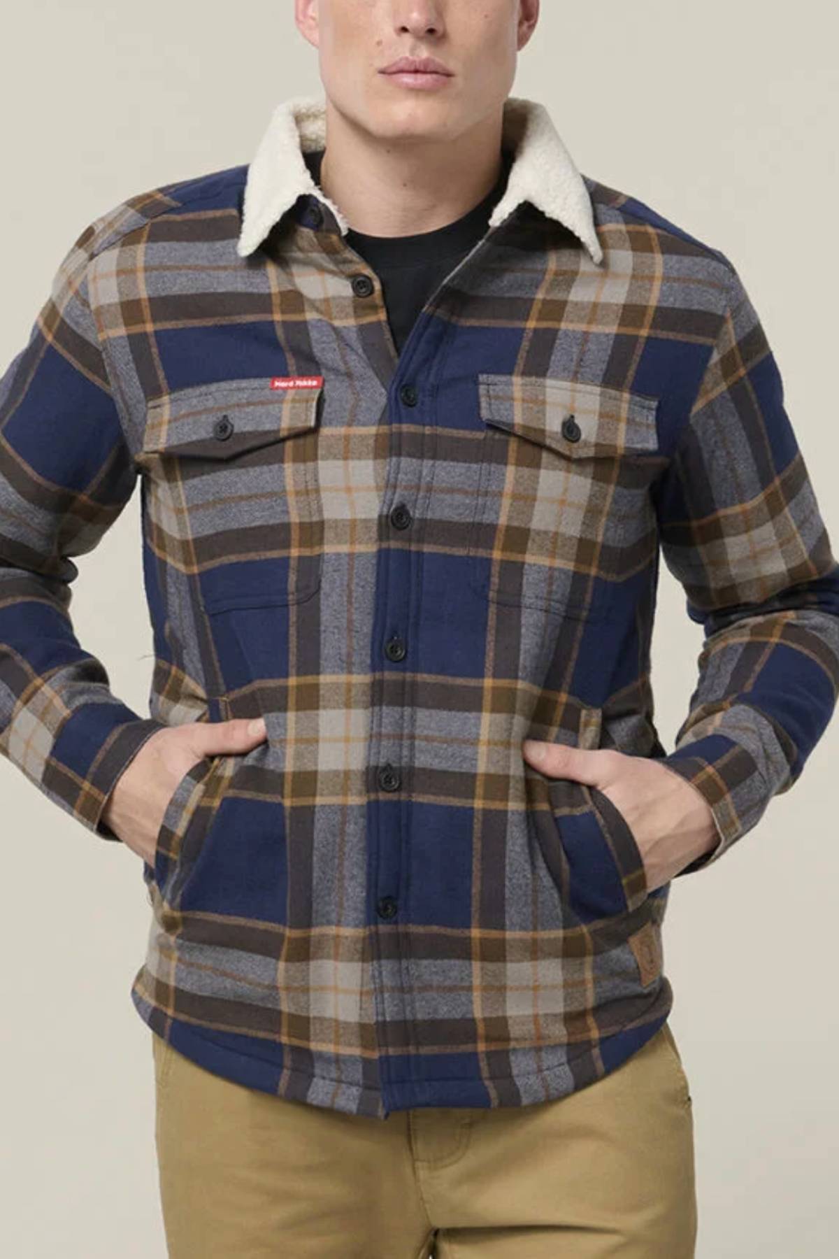 A man stands with hands in pockets, wearing a blue, brown, and gray plaid Hard Yakka Heritage Trucker Sherpa Jacket with a white sherpa collar over a black shirt and tan pants—a classic pick for any winter wardrobe.