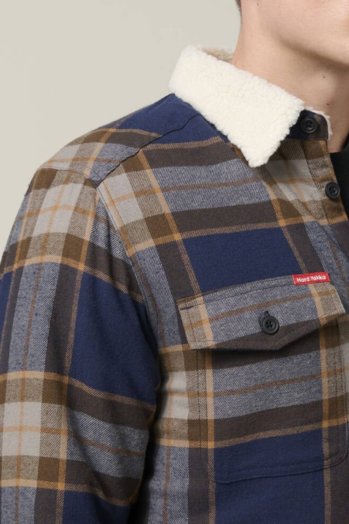 A person wears the Hard Yakka Heritage Trucker Sherpa Jacket, featuring blue and brown plaid, a white sherpa collar, and a buttoned chest pocket. Only their chest and part of their face are visible, highlighting this cozy winter staple.