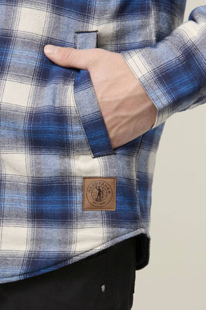 A person wears the Hard Yakka Quilted Flannelette Shacket in blue and white plaid, with one hand in the pocket. A brown patch with a circular logo is sewn near the hem above black pants, highlighting its durable style and comfort.