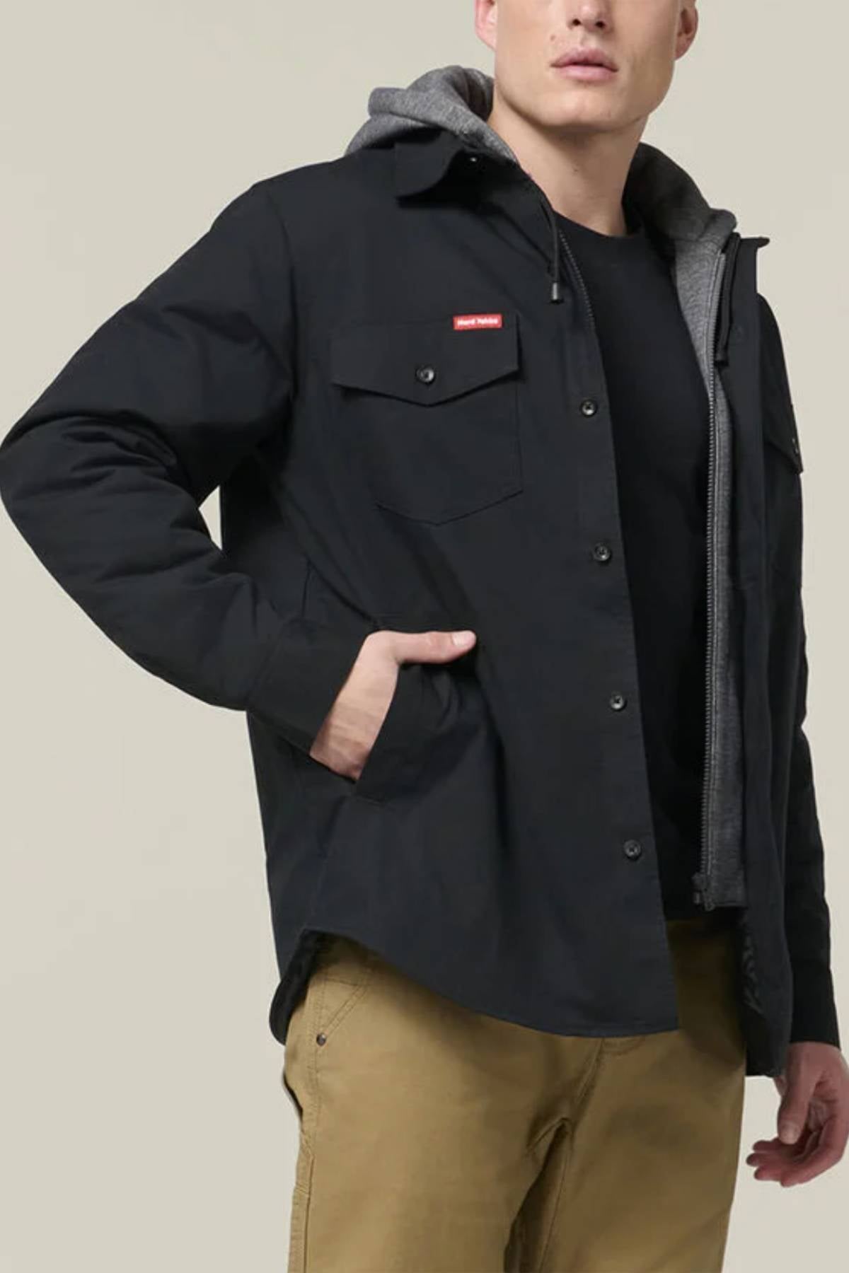 Wearing the Hard Yakka Quilted Solid Shacket in black over a grey hoodie, black tee, and tan pants, a person stands with one hand in their pocket against a plain light background.