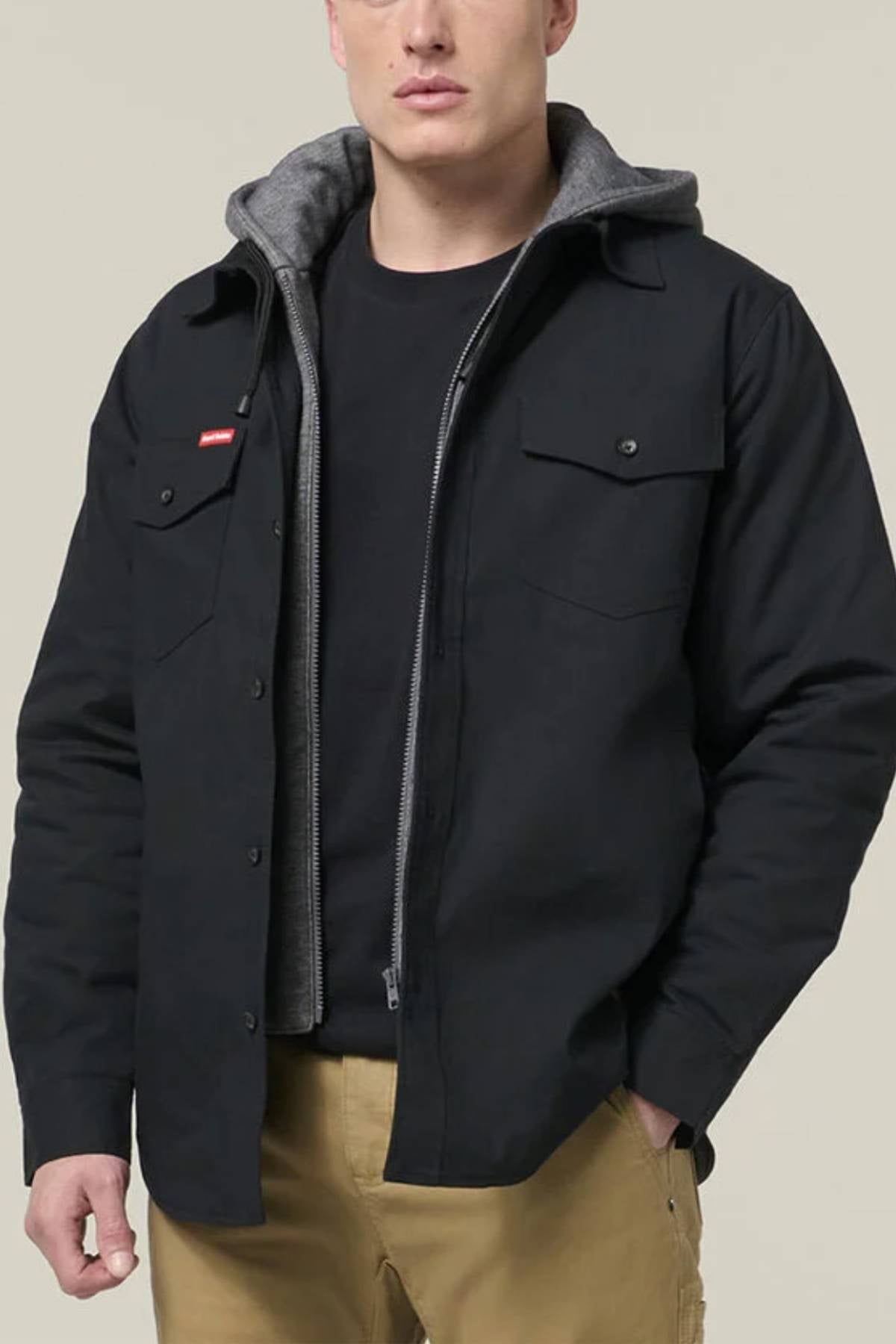 A man wearing a Hard Yakka Quilted Solid Shacket with durable Ripstop Outer Fabric over a gray zip-up hoodie, black shirt, and khaki pants stands against a plain light background.