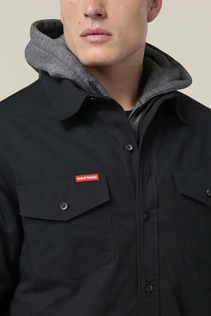 A person wearing a black Hard Yakka Quilted Solid Shacket with flap pockets over a gray hoodie. The durable ripstop shacket features a small red and white Nord Workwear label on the chest pocket. The person's face is partially visible.