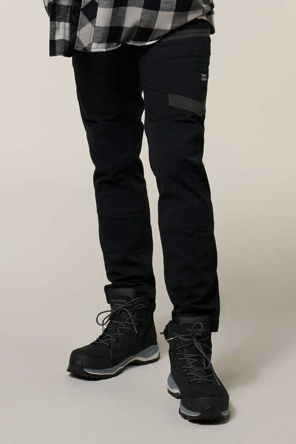 A person in black Hard Yakka Raptor Pant with Repreve ECO Ripstop Technology, black hiking boots, and a black-and-white plaid shirt stands on a light surface; the photo is cropped below the shoulders.