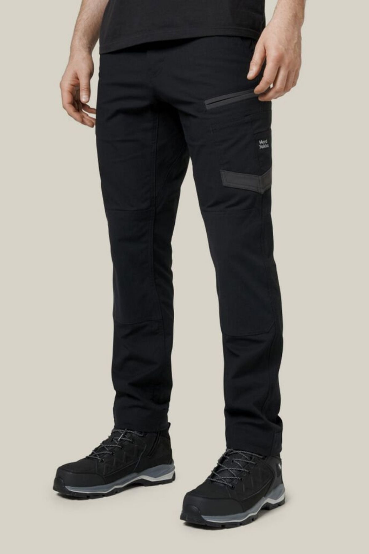 A person wears Hard Yakka Raptor Pant hiking pants with zippered pockets and black hiking shoes, standing against a plain beige background. Only the lower half of their body is visible.