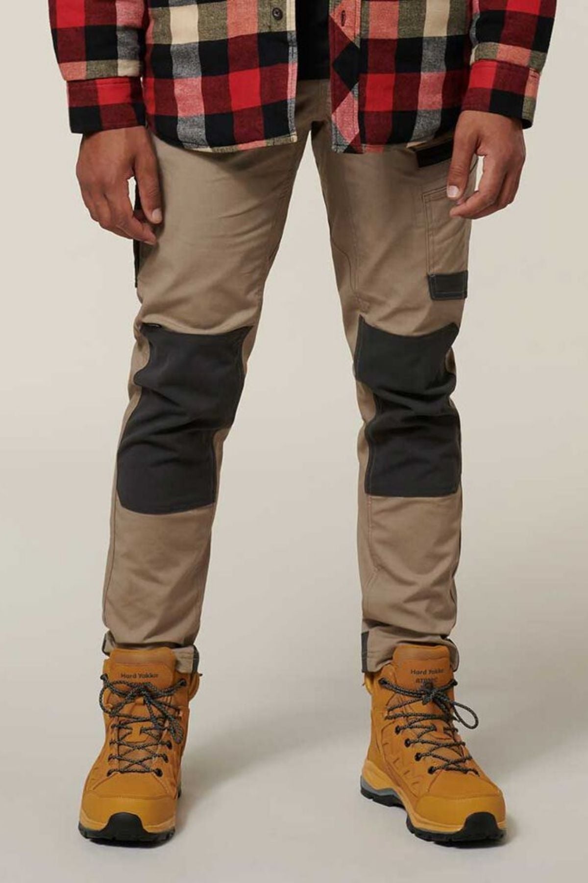 A person is standing wearing a red and black checkered jacket, tan Hard Yakka Raptor Pants with reinforced knee patches featuring Repreve ECO Ripstop Technology, and yellow hiking boots with blue laces. The background is plain and light-colored.