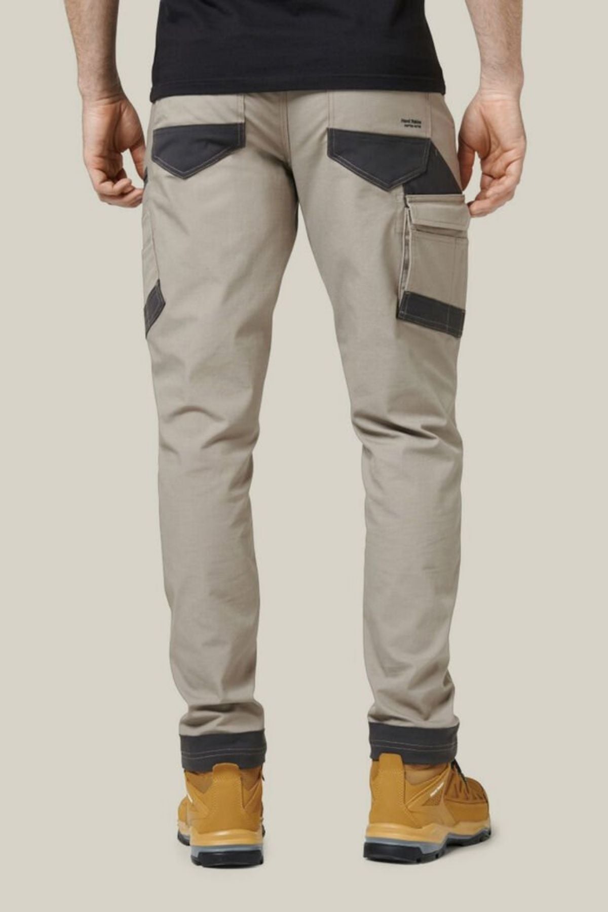 A person wearing beige Hard Yakka Raptor Pants with black pockets and mustard-yellow work boots is standing with their back facing the camera. The pants, made with Repreve ECO Ripstop Technology, feature multiple utility pockets on the sides and back. They are also wearing a black shirt.