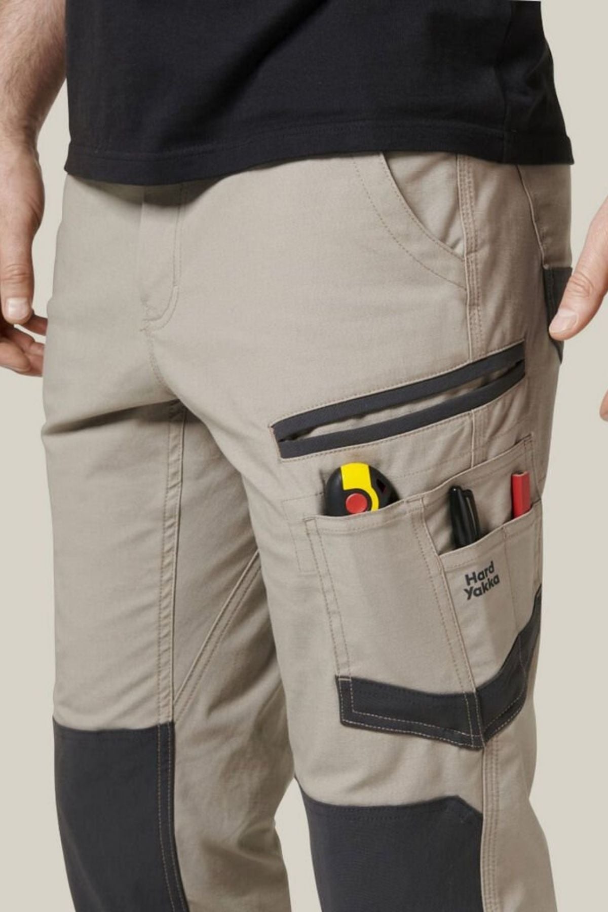 A close-up of a person wearing beige Hard Yakka Raptor Pant with black knee patches and multiple pockets. The pants feature Repreve ECO Ripstop Technology for durability. The pockets contain a utility knife, pen, and other tools, with the brand name "Hard Yakka" visible on one of them.