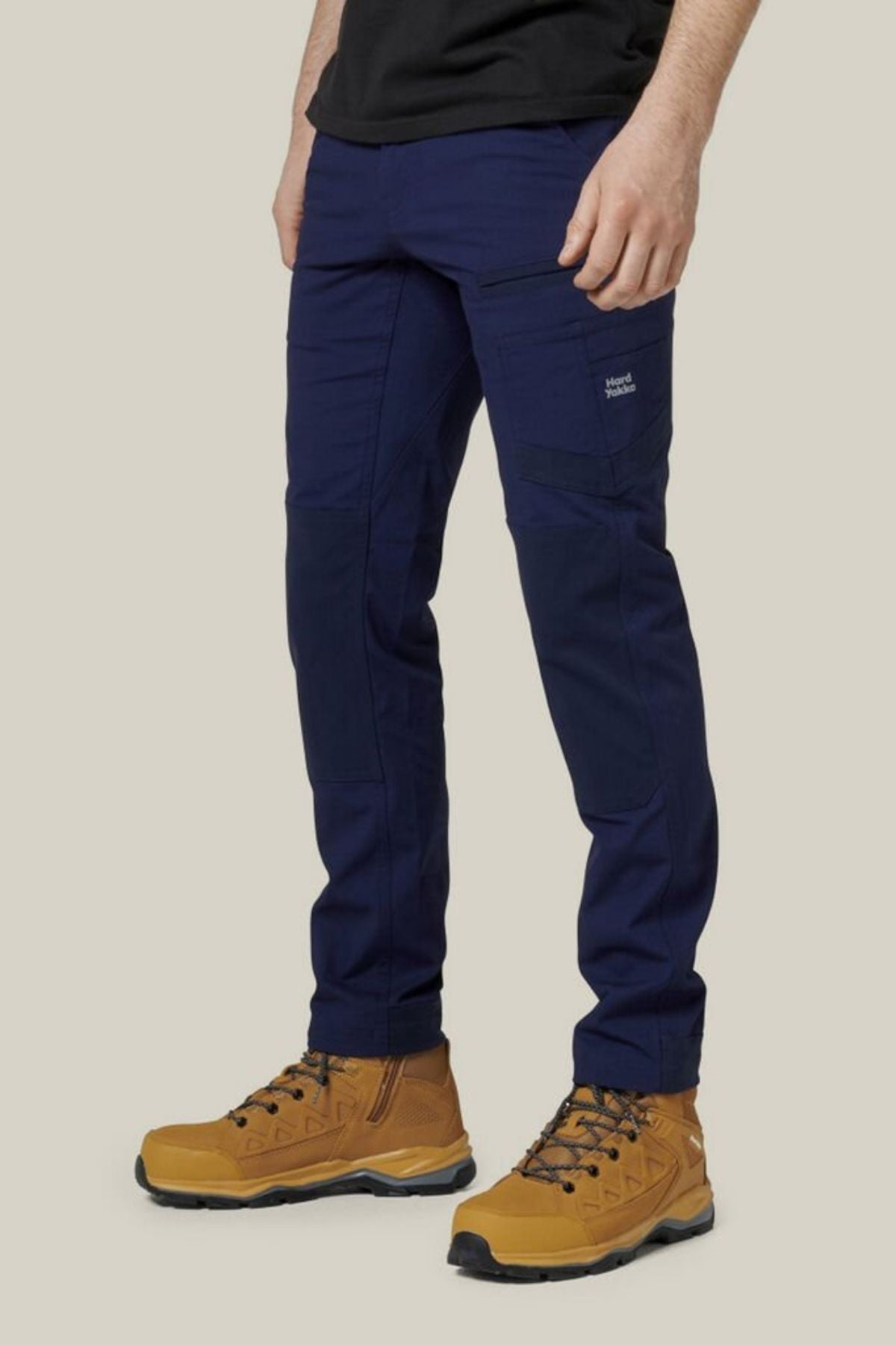 A person wearing Hard Yakka Raptor Pants in dark blue with reinforced knees and a black shirt is standing against a plain, light-colored background. They are also wearing tan hiking boots with black soles. The Hard Yakka Raptor Pant features visible pockets on the thighs and knees, showcasing ECO Ripstop Technology for durability.