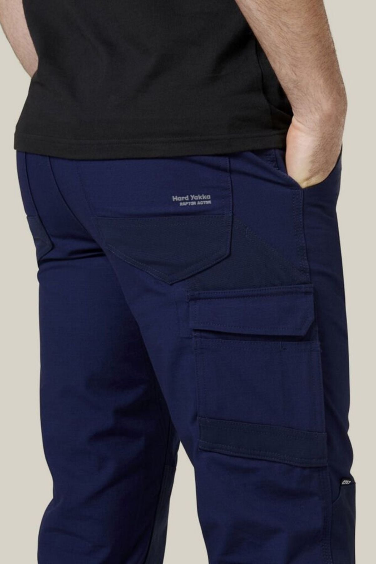 A person wearing Hard Yakka Raptor Pants in navy blue, featuring reinforced knees and a visible "Hard Yakka" logo on the back pocket. The work pant includes multiple large pockets, with one of them holding their hand. They are also wearing a black top, showcasing both style and functionality.