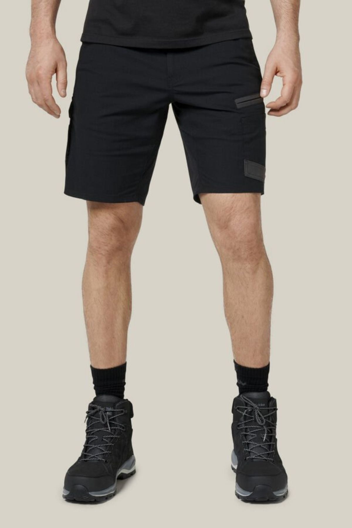 A person sports the Hard Yakka Raptor Mid Short in black, featuring Repreve ECO Ripstop Technology, paired with black crew socks and rugged hiking boots. The background is light beige. Their upper body is partially visible, showcasing a sleek black shirt.