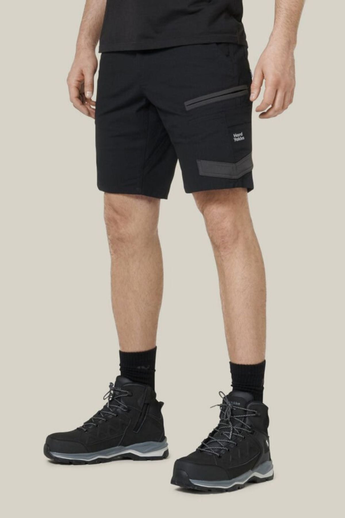 A person wearing Hard Yakka Raptor Mid Shorts and black hiking boots with gray accents stands against a plain background. The black cargo shorts feature visible pockets, an Active Fit stretch waistband, and Repreve ECO Ripstop Technology for durability, paired with black socks for a sleek look.