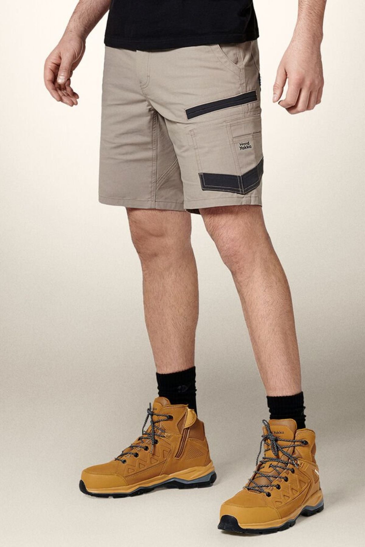 A person is wearing Hard Yakka Raptor Mid Shorts in beige, crafted from durable stretch ripstop fabric with black accents and pockets, paired with black socks and tan hiking boots. The upper part of the body is not visible, set against a plain, light-colored wall.