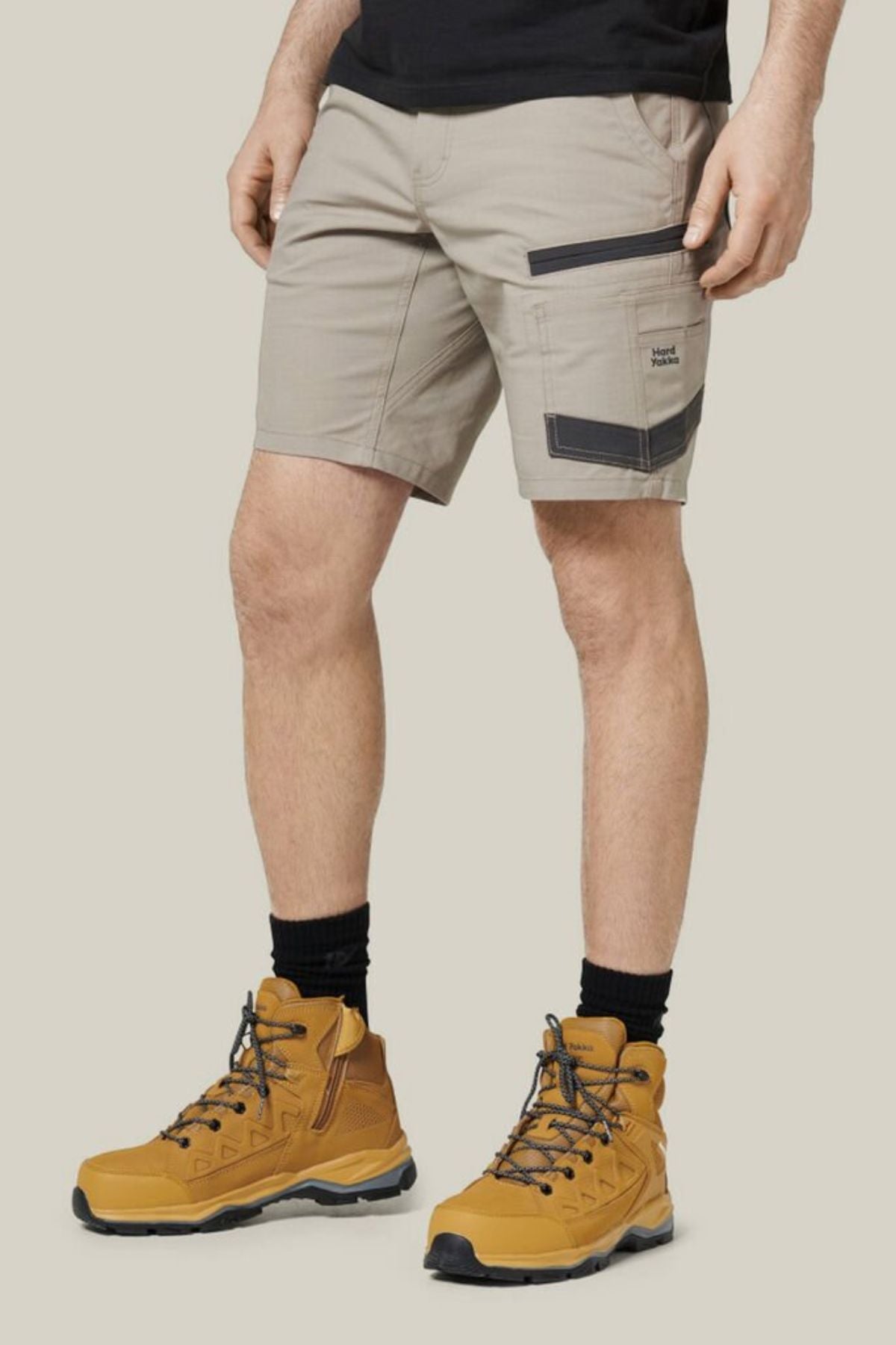 A person wearing Hard Yakka Raptor Mid Shorts in light beige, crafted from durable stretch ripstop fabric with black detailing, paired with a black t-shirt, black socks, and tan hiking boots. The person is standing against a plain, neutral background.