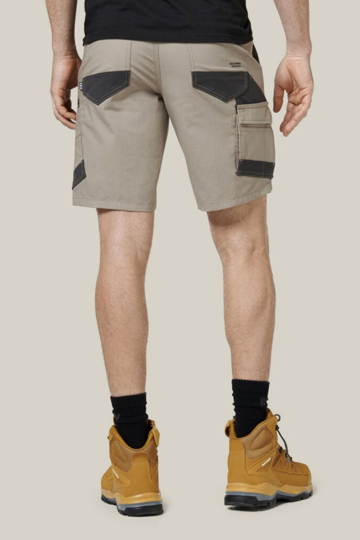 A person is standing with their back facing the camera, wearing a black T-shirt and Hard Yakka Raptor Mid Shorts in beige. They paired this outfit with black socks and tan work boots. The shorts are made from durable stretch ripstop fabric featuring black pocket detailing. The background is a plain, light color.