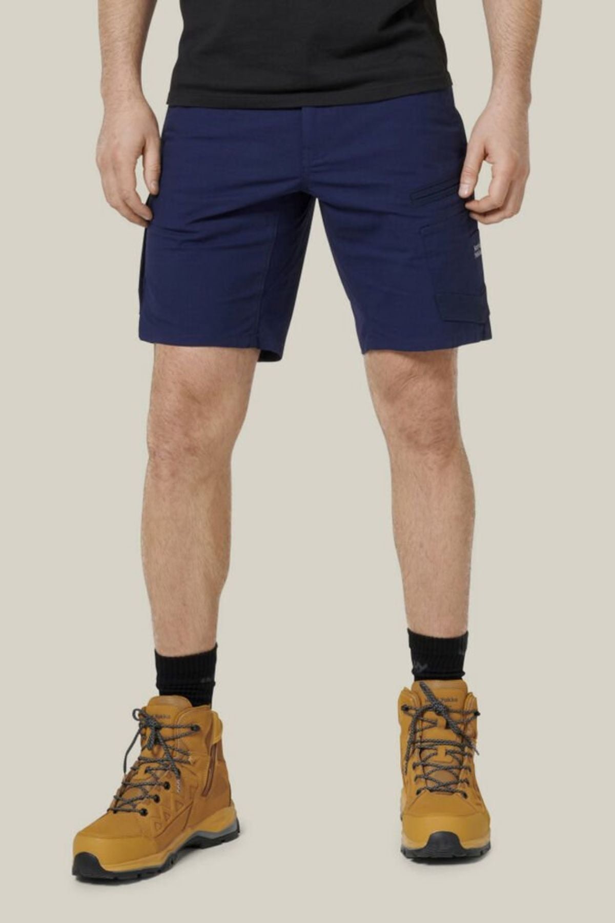 A person wearing Hard Yakka Raptor Mid Shorts in dark blue, with an Active Fit stretch waistband, a black t-shirt, black socks, and tan hiking boots stands against a plain light beige background.