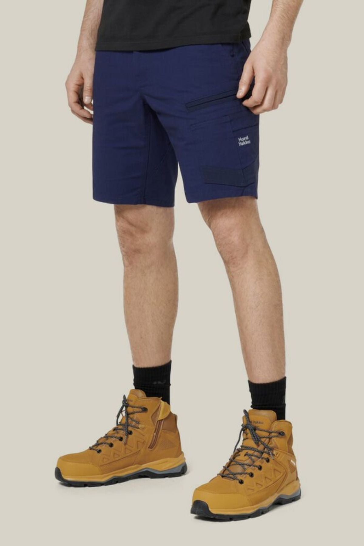 A person wearing the Hard Yakka Raptor Mid Short in navy with an Active Fit stretch waistband, black socks, and tan hiking boots stands against a beige background. The photo shows them from the waist down.