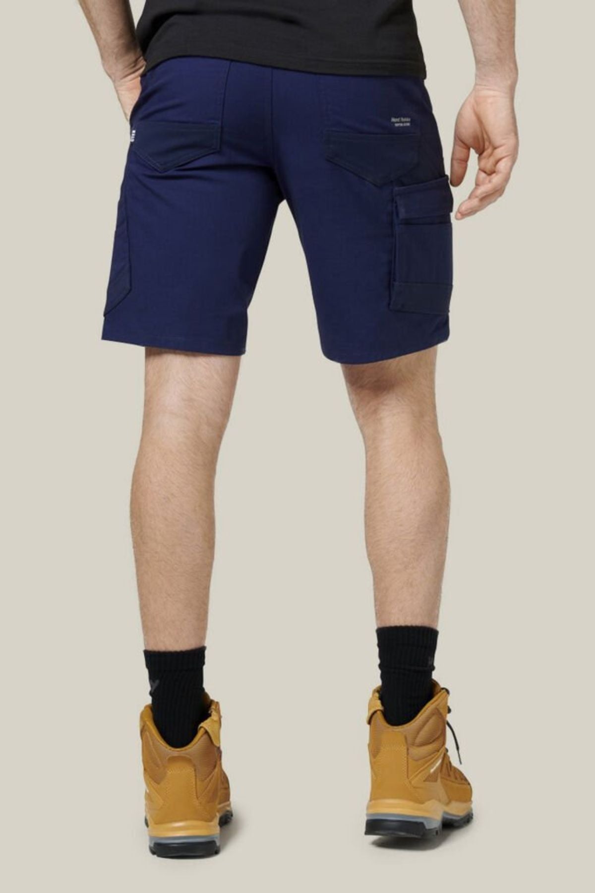 A person is shown from the back, standing against a plain light beige background, wearing Hard Yakka Raptor Mid Shorts in navy blue with an Active Fit stretch waistband, along with a black shirt, black socks, and tan boots.