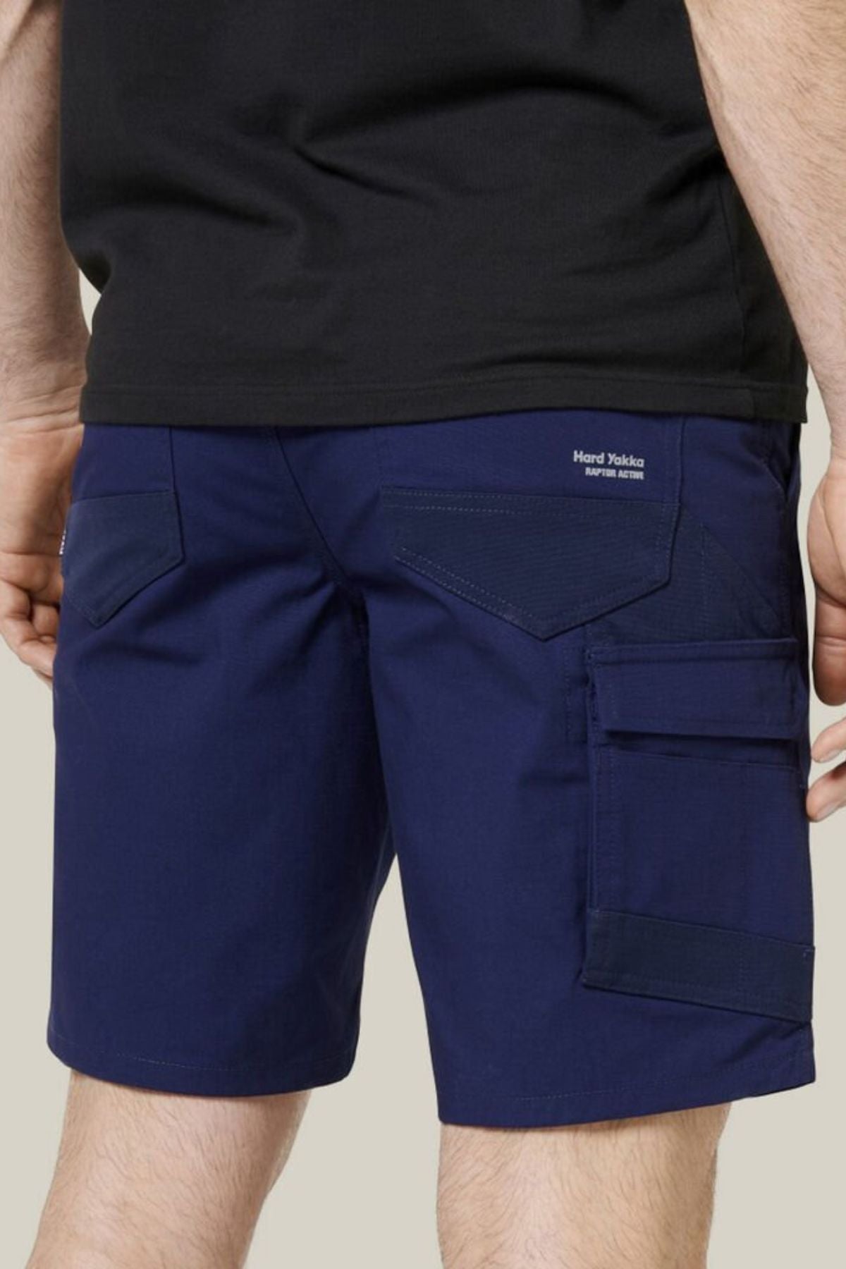 A person seen from the back wears Hard Yakka Raptor Mid Shorts in dark blue, featuring an Active Fit stretch waistband, large back pockets, and a side flap pocket, paired with a black shirt.