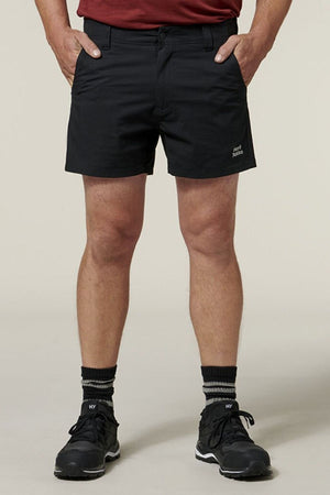 Front view of a person wearing Hard Yakka Raptor Short Shorts in black, looking comfortable and durable with hands in pockets, complemented by black hiking boots, white socks with black stripes, and a red shirt. The background is plain and light-colored.