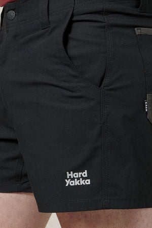 Close-up of a person wearing dark-colored Hard Yakka Raptor Short Shorts featuring multiple pockets. The brand name "Hard Yakka" is prominently displayed in white on the lower part of the shorts. These comfortable, hard-wearing shorts also feature an Active Fit waistband for added comfort.