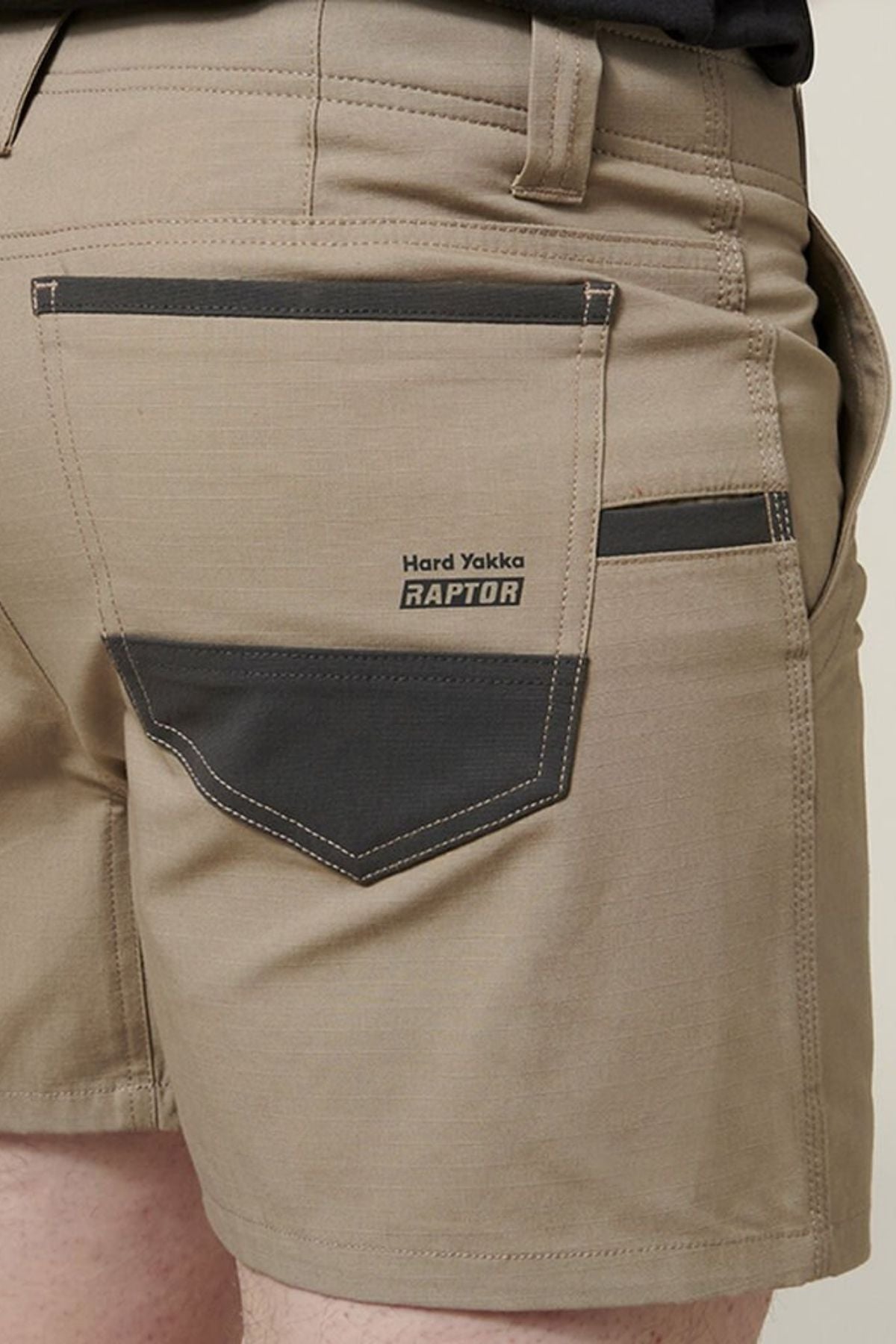 A person wears Hard Yakka Raptor Short Shorts in beige with dark gray accents. The Active Fit waistband offers comfort, while a pocket showcases "Hard Yakka RAPTOR." Made with durable Sustainable REPREVE® ECO Ripstop Technology, these shorts are both tough and eco-friendly.