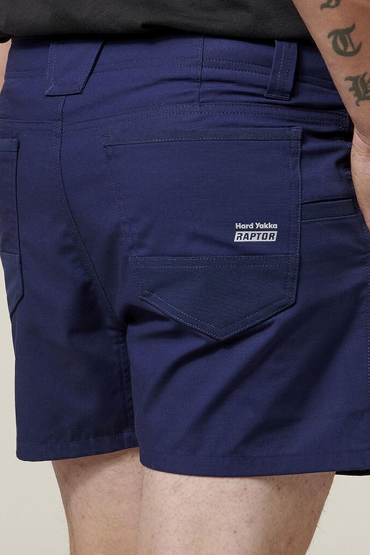Close-up of a person wearing dark blue "Hard Yakka Raptor Short Shorts," featuring multiple utility pockets for practicality. Made from sustainable fabric, these comfortable shorts are perfect for daily wear. The individual, sporting a tattoo on their arm, stands against a plain background.