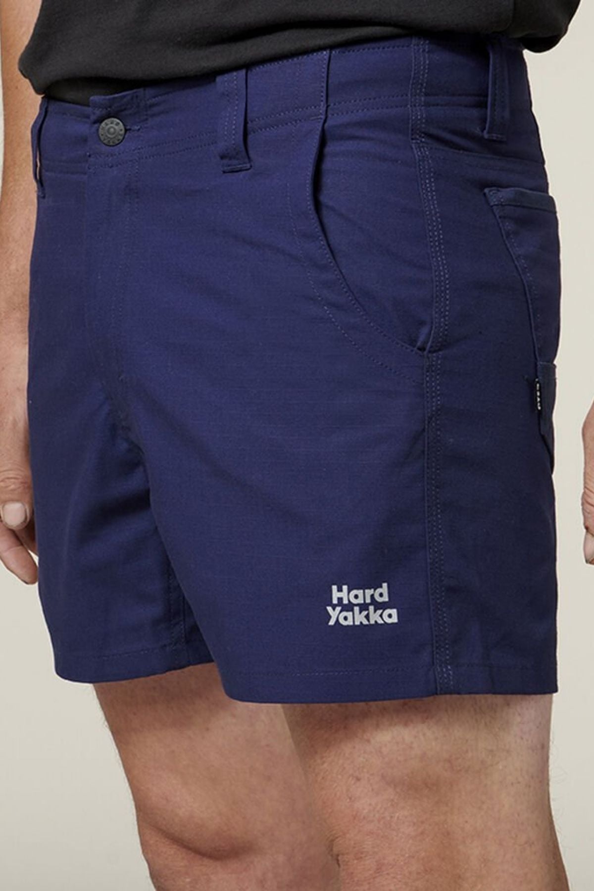 Close-up of a person wearing dark blue Hard Yakka Raptor Short Shorts. These comfortable shorts feature front utility pockets and a button closure, with the Hard Yakka logo printed in white on the lower left side. The person is wearing a black shirt tucked into the shorts.