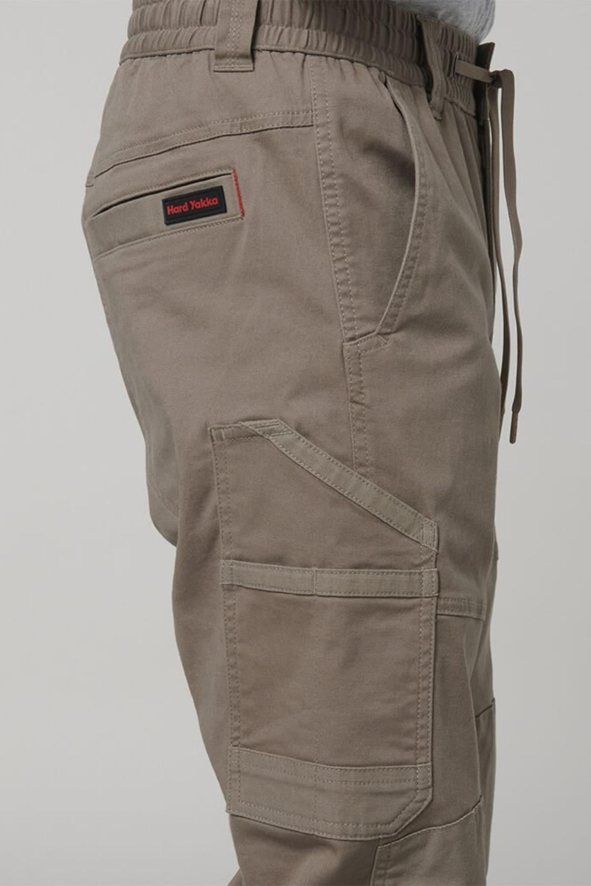 Side view of a person wearing Hard Yakka Tough Maxx Pant cargo pants featuring multiple pockets and a drawstring waistband. A red and black "Hard Yakka" label is visible above the left thigh pocket.