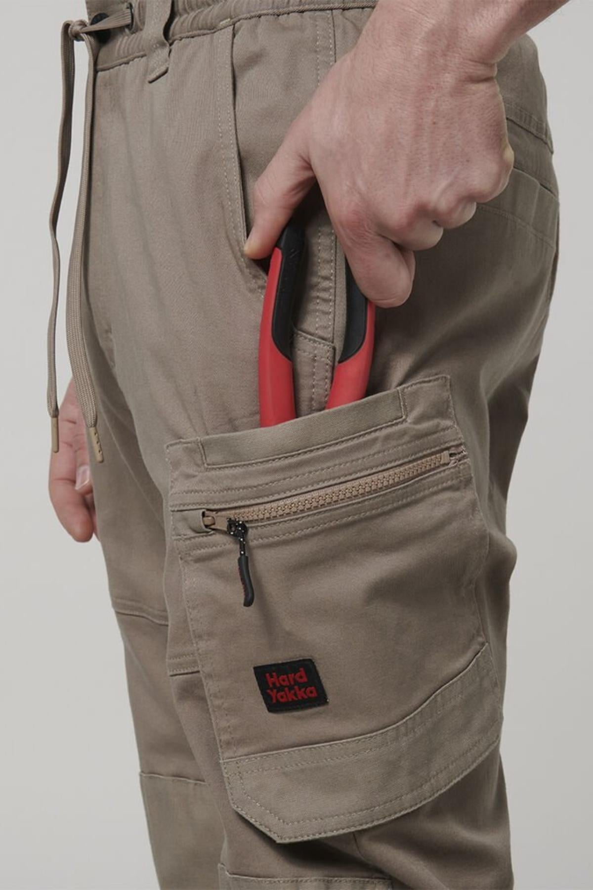 Wearing Hard Yakka Tough Maxx Pants in beige with multiple pockets, a person puts red-handled pliers into a zippered side pocket featuring the Hard Yakka label.