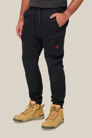 A person stands against a light beige background wearing Hard Yakka Toughmaxx Cuffed Pants with a drawstring waist and zippered pocket, paired with tan lace-up work boots, highlighting the pants' reinforced durability.