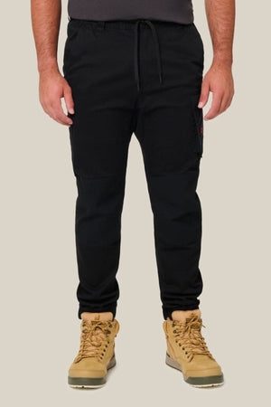 A person stands against a plain beige background, shown from the shoulders down, wearing Hard Yakka Toughmaxx Cuffed Pants in black—renowned for reinforced durability—and tan lace-up boots.