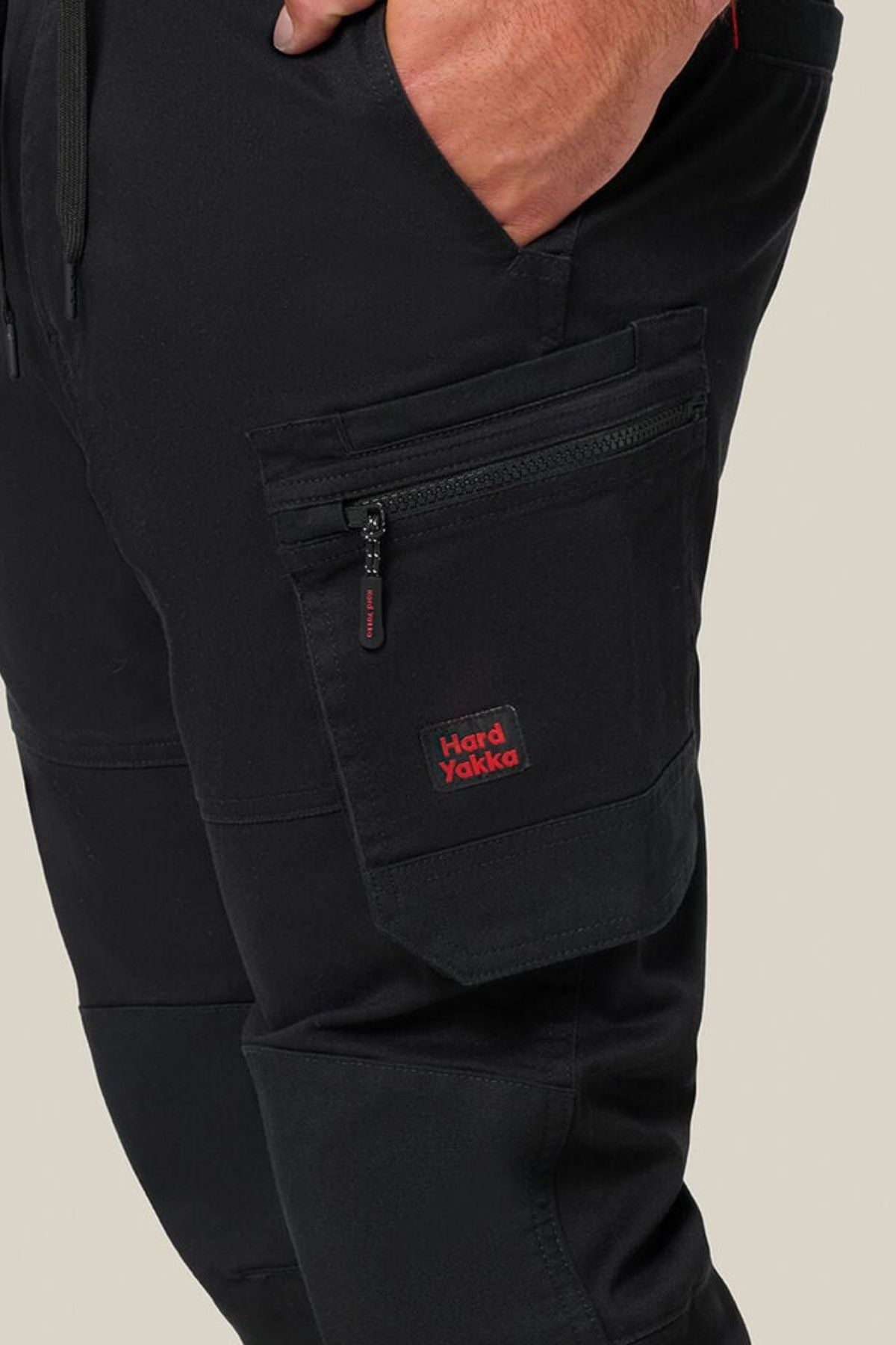 Close-up of a hand in pocket, wearing black Hard Yakka Toughmaxx Cuffed Pant with reinforced durability and a zippered side pocket. The red and black Hard Yakka logo is visible on the pocket. Background is plain beige.