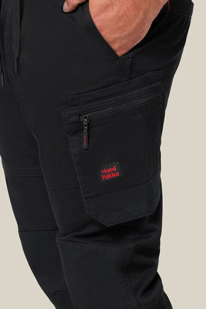 Close-up of a hand in pocket, wearing black Hard Yakka Toughmaxx Cuffed Pant with reinforced durability and a zippered side pocket. The red and black Hard Yakka logo is visible on the pocket. Background is plain beige.