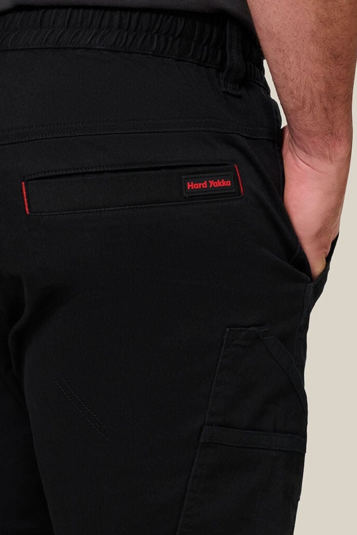 Close-up of a person wearing Hard Yakka Toughmaxx Cuffed Pant in black, featuring reinforced durability, elastic waistband, and a back pocket with red-stitched “Hard Yakka” label, one hand casually placed in the pocket.