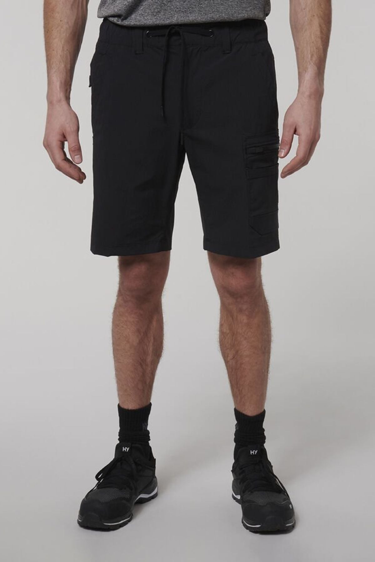 A person models Hard Yakka X Range Mid Shorts in black Cordura nylon, paired with a gray shirt, black socks, and black athletic shoes, standing against a plain light background. The image is cropped from the shoulders down.