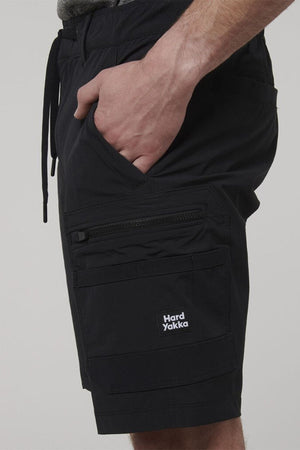 A person wearing Hard Yakka X Range Mid Shorts with their hand in a pocket. These black work shorts feature multiple pockets, a zipper, drawstring waist, and a visible "Hard Yakka" logo—ideal for tough tasks.