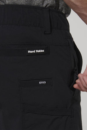 Close-up of a person wearing Hard Yakka X Range Mid Short in black, made from durable workwear fabric. Zippered and flap pockets feature "Hard Yakka" and "CTE3" labels, with a hand resting near the side pocket.