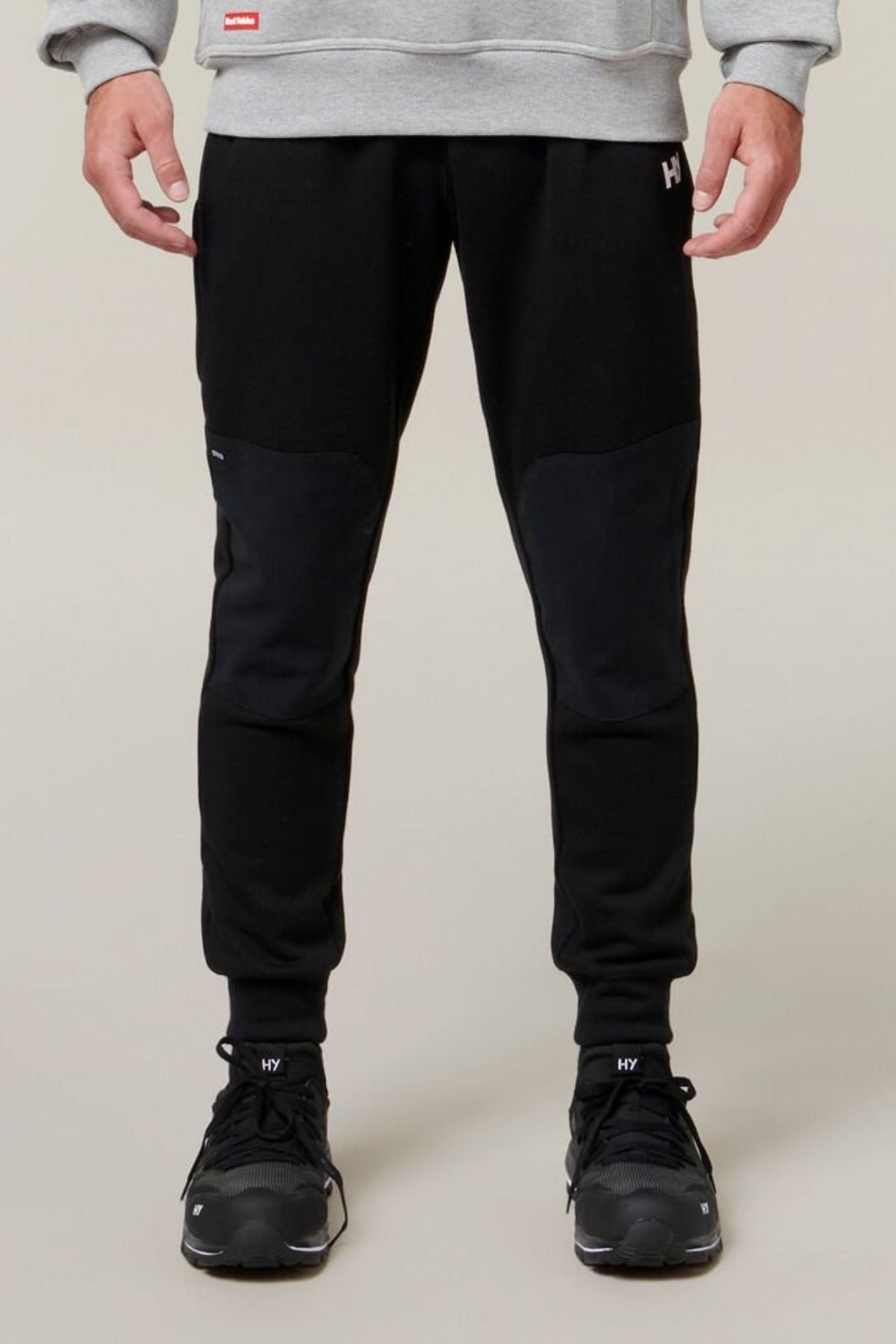 A person stands facing forward, wearing a grey sweatshirt, Hard Yakka Xtreme Jogger black track pants with reinforced knee patches, and black sneakers. Only the lower half of the body is visible against a plain background.
